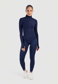 Marineblauwe aansluitende jas met ritssluiting, lange mouwen en duimgaten, gecombineerd met bijpassende legging. Soepele, rekbare stof voor comfort en flexibiliteit.