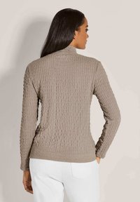 Frau mit langen dunklen Haaren, die ein beigefarbenes, strukturiertes Langarmoberteil und weiße Hosen trägt. Sie steht mit einer Hand in der Tasche und schaut weg.