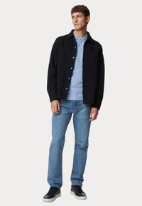Marineblaue Jacke mit Knopfleiste und zwei Brusttaschen über einem hellblauen Poloshirt, kombiniert mit hellen Jeans und dunklen Sneakers.