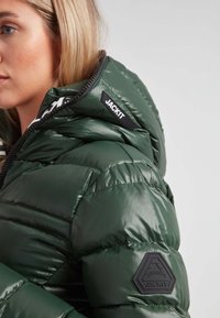 Chaqueta de abrigo verde con textura brillante, cierre de cremallera y parches del logo en la capucha y la manga. Detalles acolchados notables en la tela.