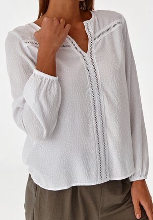Blusa bianca a maniche lunghe in tessuto texturizzato, con scollo a V e dettagli in pizzo sul davanti. Polsini elastici e vestibilità ampia.