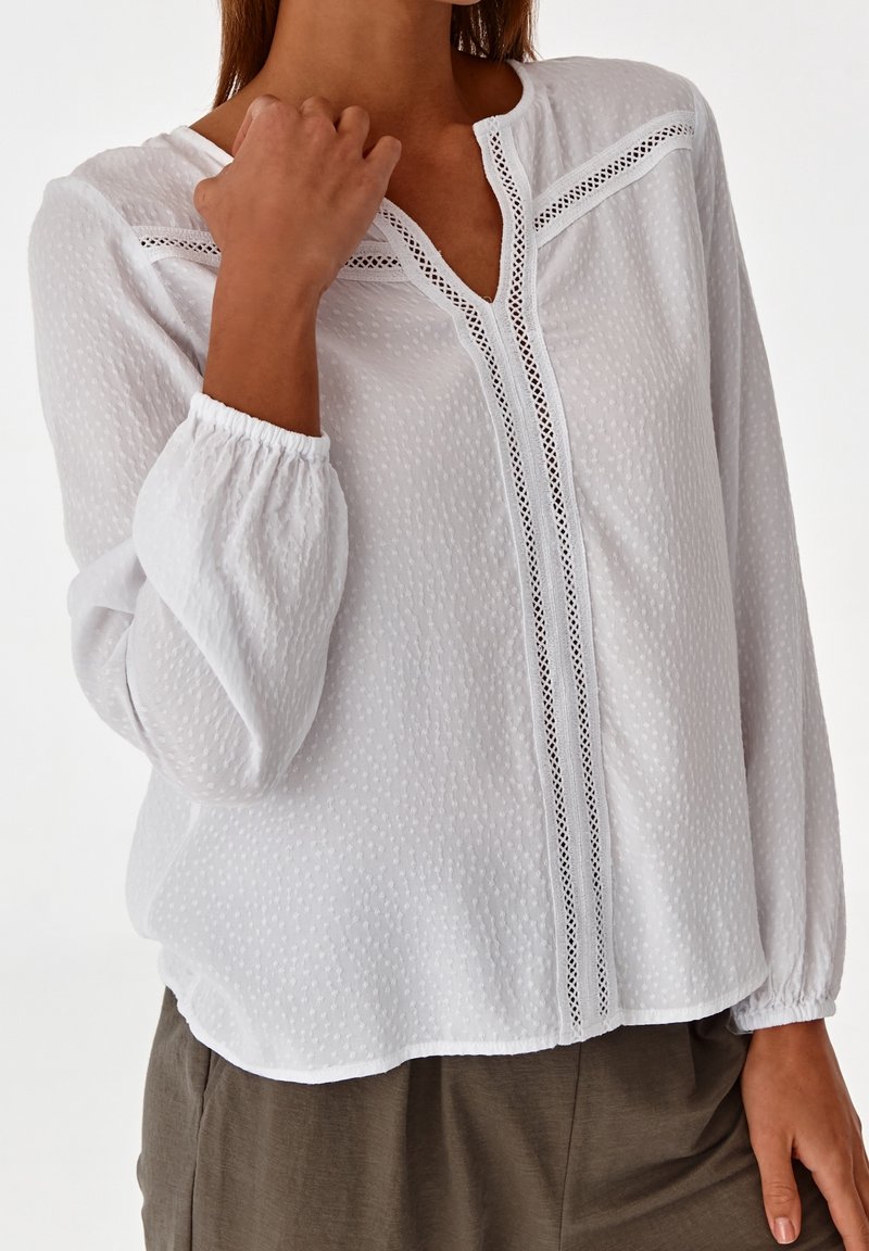 Blusa bianca a maniche lunghe in tessuto texturizzato, con scollo a V e dettagli in pizzo sul davanti. Polsini elastici e vestibilità ampia.