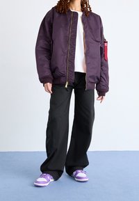 Lila bomberjacka med en gul dragkedja, ribbade ärmmuddar och en röd etikett. Svarta vida byxor och sneakers med lila detaljer fulländar utseendet.