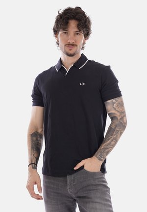 Uomo con capelli ricci e tatuaggi su entrambe le braccia, indossa una polo nera e jeans grigi, in piedi con una mano in tasca.