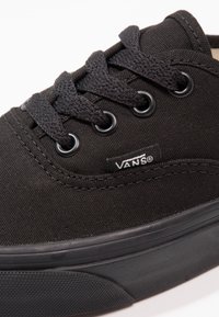 Gros plan d'une chaussure Vans noire avec une tige en tissu, des lacets noirs passant à travers des œillets en métal, et une étiquette avec le logo Vans visible sur le côté.