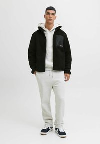Giacca in pile nera con tasca con zip, felpa grigia sotto, pantaloni della tuta grigi e sneakers blu con lacci bianchi e suola beige; modello in piedi.