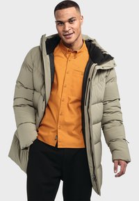Puffermantel in hellem Olivgrün mit einem gesteppte Design, verstellbarer Kapuze und schwarzem Innenfutter. Über einem orangefarbenen Hemd mit Knopfverschluss getragen.