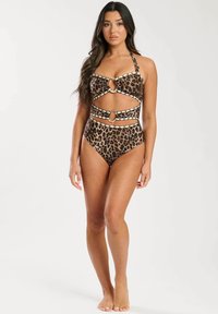 Kvinde står barfodet og bærer en one-piece badedragt med leopardmønster og ringdetaljer på en ensfarvet lys baggrund.