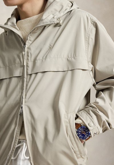 Homme portant une veste beige zippée et un pull en maille crème avec une montre au cadran bleu au poignet gauche, main dans la poche de la veste.