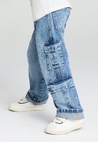 Niebieskie dżinsowe spodnie cargo z wieloma kieszeniami, luźny krój i podwinięte mankiety. Noszone z białymi sneakersami z detalami w postaci paska i granatowymi akcentami.