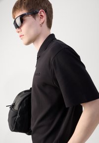 Svart polo-skjorte med glidelåskrage, med merket "HUGO" på brystet, kombinert med en svart teksturert ryggsekk og solbriller.