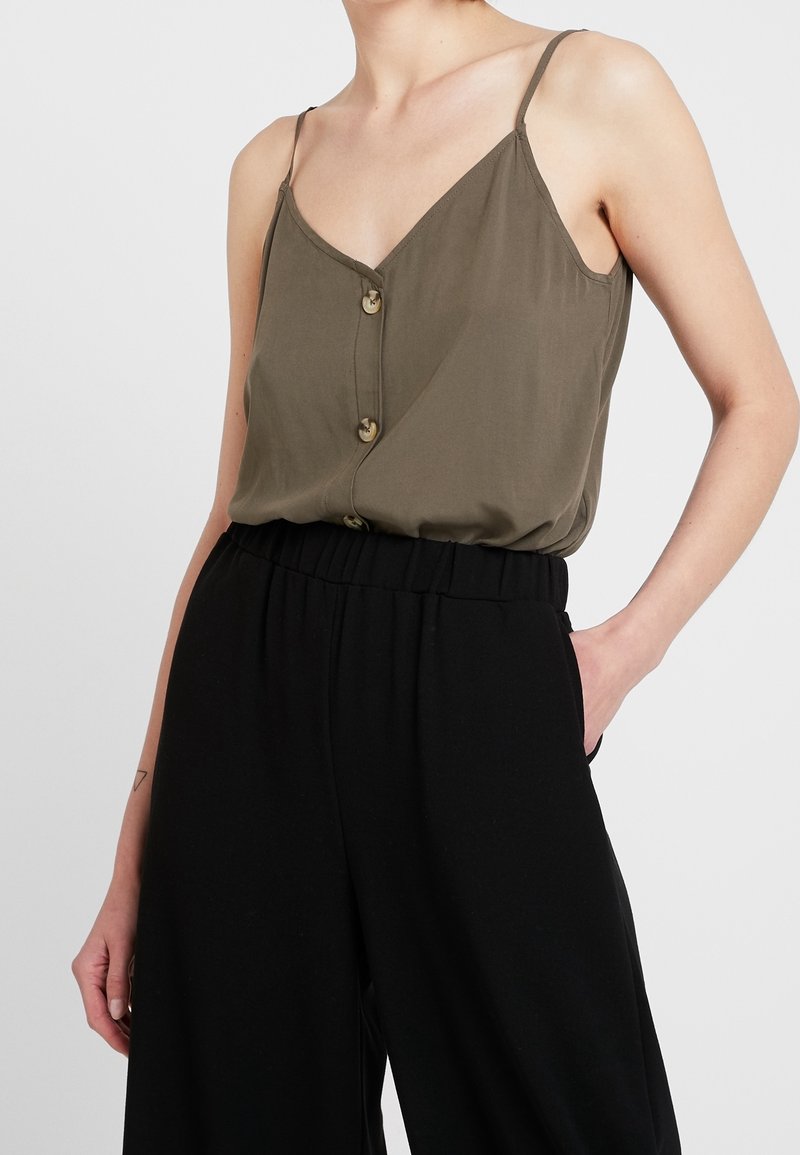 Top sans manches vert olive avec un décolleté en V et de petits boutons, associé à un pantalon noir ample taille haute, doté de poches latérales.