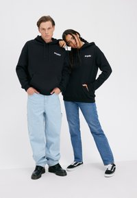 Kaks inimest seisavad kõrvuti, kandes musti kapuutsega pusse, millel on tekst "MEMORIZE", siniseid teksapükse ja vabas stiilis musti kingi tavalise tausta ees.