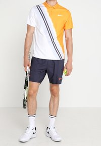 Homme debout portant un polo orange et blanc, un short bleu marine, des baskets blanches, tenant une raquette de tennis dans la main gauche et une balle de tennis dans la main droite.