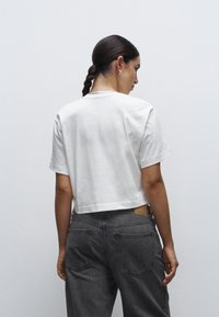 Camiseta blanca cortada de algodón, con mangas cortas. Combinada con jeans gris de tiro alto, con bolsillos traseros y una etiqueta texturizada.