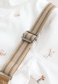 Beige elastic suspenders with dark striped accents, attached to a white shirt featuring small, colorful airplane patterns.