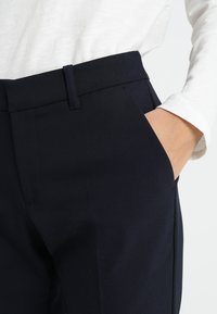 Navy blue tailored trousers featuring a smooth texture, side pockets, and a classic waistband design. Styled with a white long-sleeve top.