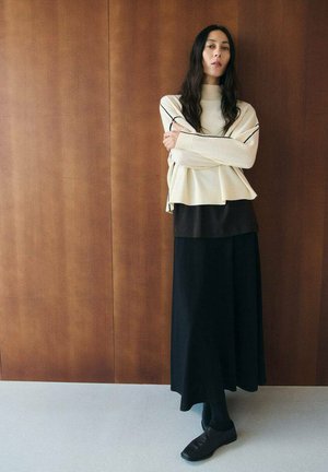 Donna con capelli lunghi e scuri, in piedi contro un muro di legno, indossa un maglione a collo alto crema con dettagli neri, gonna nera, scarpe nere.