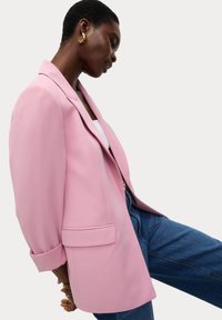 Blazer rose clair à la coupe décontractée, avec un col cranté, une poche avant simple et des manches retroussées, associé à un jean denim foncé.