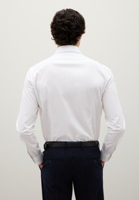 Uomo con capelli ricci scuri che indossa una camicia bianca infilata in pantaloni a righe navy con cintura nera, mani nelle tasche, di spalle.