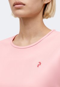 Κοντινό πλάνο ενός ατόμου που φοράει ανοιχτό ροζ πουκάμισο με μικρό κόκκινο γεωμετρικό λογότυπο στο στήθος, με κοντά ξανθά μαλλιά ορατά.