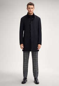Giovane uomo con un cappotto nero con bottoni, pantaloni a quadri grigi e stivali di pelle neri, in piedi su uno sfondo chiaro e semplice.