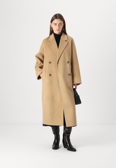 Cappotto in lana color cammello a doppio petto con colletto a revers, tasche laterali e vestibilità rilassata. Abbinato a stivali neri fino al ginocchio.