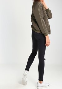 Bomber bunda olivové barvy s prošíváním a žebrovanými manžetami, kombinovaná s černými skinny džínami a bílými šněrovacími teniskami. Jasné, texturované detaily jsou viditelné.