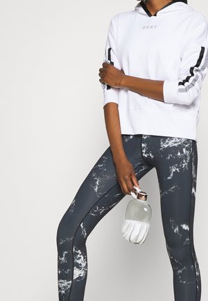 Osoba ubrana w biały kaptur z czarnymi paskami na rękawach oraz czarno-białe legginsy w marmurkowy wzór, trzymająca przezroczystą butelkę na wodę z marmurkowym pokrowcem.