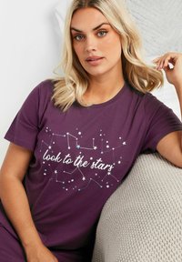 Lila bomull t-shirt med ett stjärnbildsgrafik och texten "titta mot stjärnorna" i vitt och pastellfärger. Korta ärmar, avslappnad passform.