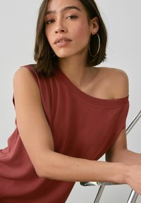 Top de color óxido, sin mangas, en una tela suave, con un corte holgado. Modelo con el pelo a la altura de los hombros se recuesta contra una silla.