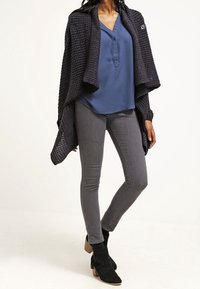 Mörkgrå stickad cardigan över en blå blus, i kombination med grå smala jeans och svarta ankelstövlar med bruna klackar.