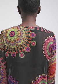 Haut en tissu noir transparent avec des motifs floraux colorés en vert, violet, rose et orange ; col rond et manches longues.