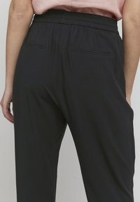 Schwarze Hose mit elastischem Bund, zwei Gesäßtaschen, glatte Textur und entspannte Passform, von hinten gezeigt, mit einem sichtbaren pinken Oberteil darüber.