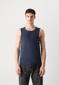 Mørk marineblå ermeløs tank topp med rund hals og subtil logo detalj. Laget av myk, lett bomullsstoff. Bært med mørke jeans.