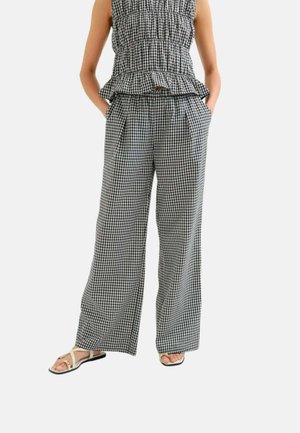 Mujer que lleva pantalones anchos de cuadros en blanco y negro y un top sin mangas a juego, con las manos en los bolsillos y sandalias blancas.