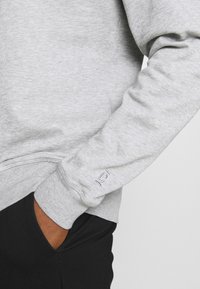 Sweat-shirt gris avec poignets et ourlet côtelés, fabriqué en tissu doux. Présente un petit logo brodé sur la manche. Pantalon noir visible.