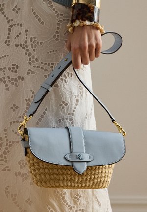 Mano sosteniendo un bolso tejido de paja y cuero azul con herrajes dorados, combinado con un vestido de encaje y pulseras de concha y carey.