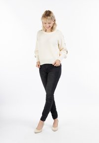 Krém színű pulóver bordázott aljjal és puffos ujjakkal, sötét, slim fit farmernadrággal és világos színű magassarkú cipővel egy sima háttér előtt.