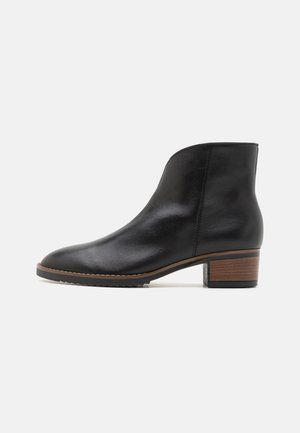 Juodas odinis kulkšninis botas su apvalia pėda, žemu sukrautu mediniu kulnu ir lygia tekstūra. Pasižymi minimaliu siuvimo darbu ir siaurėjimu.