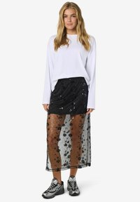 Gonna nera con pailette, con una sovrapposizione di pizzo floreale trasparente, abbinata a una maglietta bianca a maniche lunghe e scarpe da ginnastica grigie.