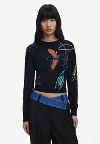 Pull noir en maille avec des broderies florales colorées en rouge, bleu et jaune, associé à un pantalon noir taille haute et une ceinture en denim.