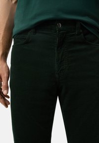 Ciemnozielone spodnie kordowe o teksturowanym wykończeniu, z zapinaniem na guziki z przodu i slim fit. Widać kieszeń boczną.