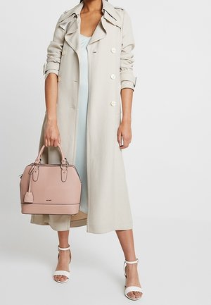 Kobieta ubrana w beżowy płaszcz trencz na jasnej sukience, trzyma różową torebkę w odcieniu brzoskwini oraz nosi białe, otwarte sandały na obcasie z paskiem wokół kostki.