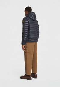Giacca a vento blu navy con cappuccio, caratterizzata da una texture liscia e lucida e un design trapuntato orizzontale. Indossata con pantaloni cargo marroni e scarpe scure.