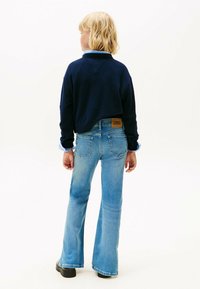 Suéter recortado azul marino con cuello acanalado, usado sobre una camisa de cuello azul claro, combinado con jeans azules acampanados que presentan una etiqueta de marca de cuero.