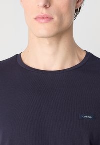 Σκοτεινό μπλε μπλουζάκι φτιαγμένο από μαλακό βαμβάκι, με λαιμόκοψη σε σχήμα crew και ένα μικρό ορθογώνιο λογότυπο Calvin Klein στο αριστερό στήθος.