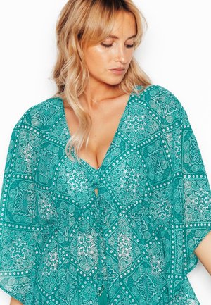 Femme blonde regardant vers le bas, portant un chemisier bleu sarcelle à motifs, col en V, manches larges et un lien à l'avant avec des pompons.