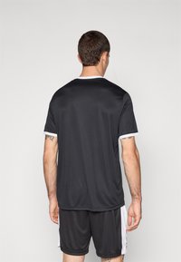 Schwarzes Sporttrikot mit kurzen Ärmeln und einem weißen Kragen, aus glattem Stoff gefertigt. Getragen mit passenden schwarzen Shorts; sichtbare Tattoos am Arm.