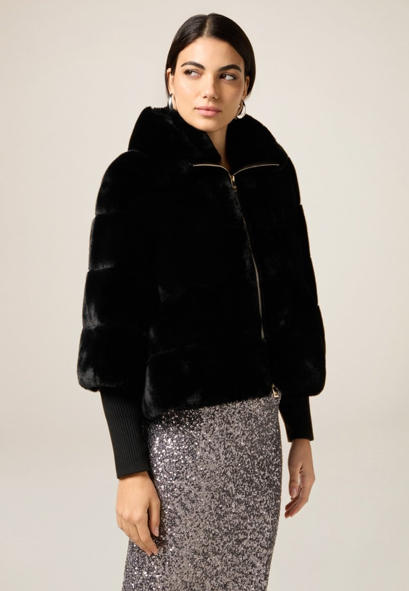 Mujer vestida con una chaqueta de pelo sintético negro con cremallera y una falda de lentejuelas plateadas, mirando de lado contra un fondo sencillo.